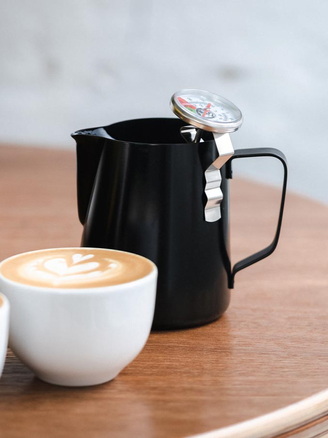 Rhino Milchkännchen mit Milchthermometer neben einer Tasse Cappuccino